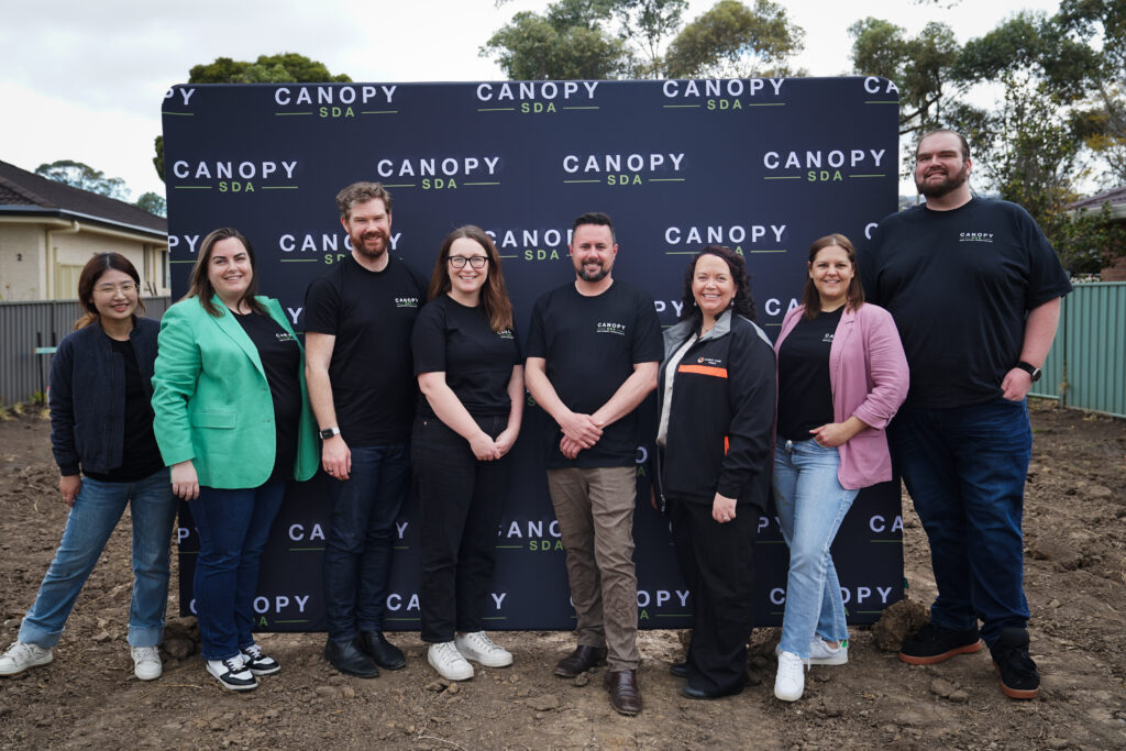 Group of 4 women and 4 men, standing on dirt in front of a big black banner that says 'Canopy SDA'. Group are all smiling.